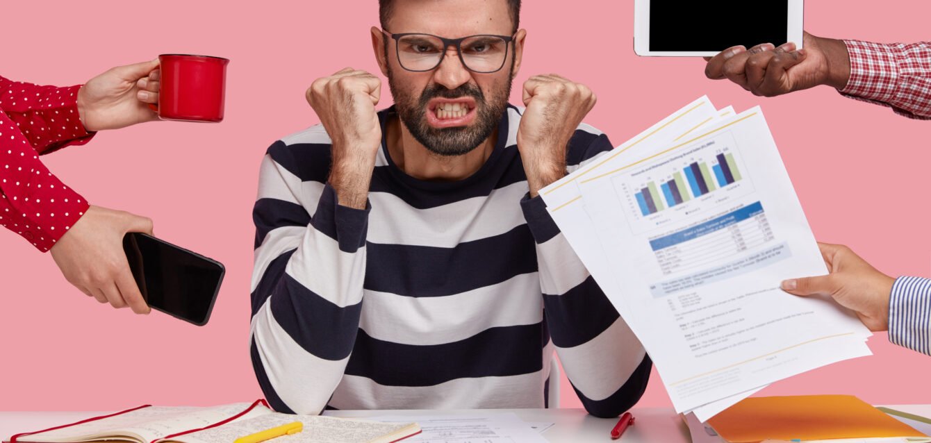 Outraged angry man has thick stubble, clenches fists in anger, unrecognizable people stretch hands to him with papers, mobile phone, notepad and mug, feels irritated with everybody, asks not bother