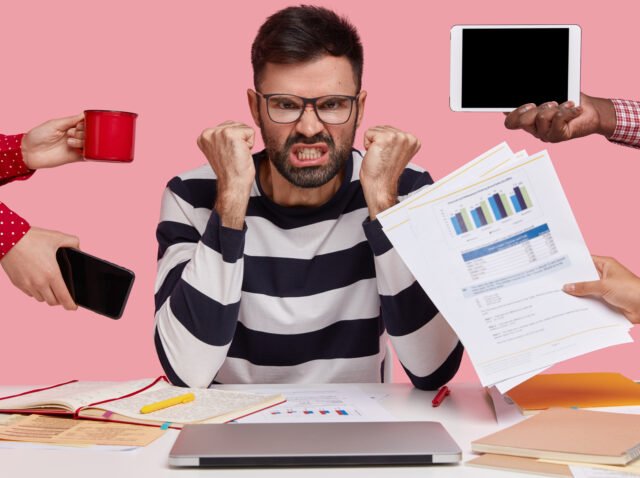 Outraged angry man has thick stubble, clenches fists in anger, unrecognizable people stretch hands to him with papers, mobile phone, notepad and mug, feels irritated with everybody, asks not bother